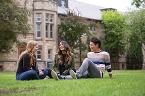 Two female and one male student sitting on the grass in front of the University of Saskatchewan, talking to each other and laughing.
