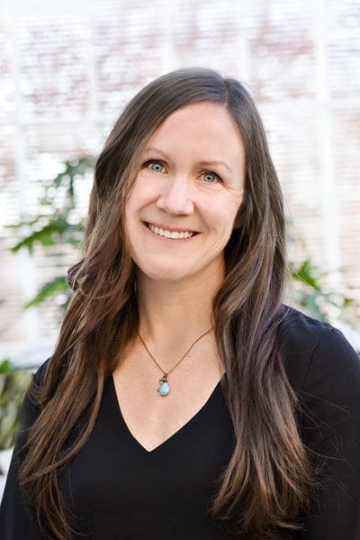 Person wearing a black V-neck top and a delicate gold necklace with a turquoise pendant. The background features softly lit windows with a blurred view of greenery.