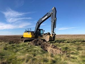A yellow and black excavator digs a deep trench in a grassy field under a bright, partly cloudy sky.