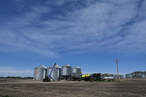 A row of large metal grain silos stands on a flat rural field under a mostly clear sky. In front of the silos, various farming equipment including tractors and machinery are arranged