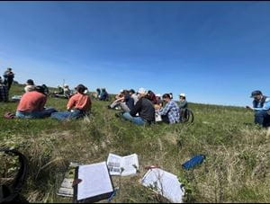Attendees at a field tour event People sitting in a grassy field during an outdoor workshop, with several participants spaced out on the hillside taking notes