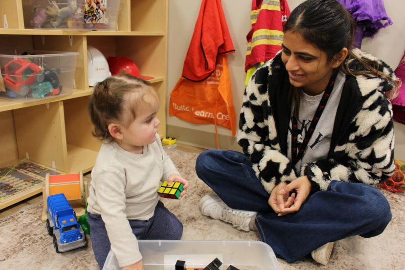 A young child is holding a rubrics cube and staring at the early childhood educator sitting beside her. They are both engaged with each other and the task at hand. 
