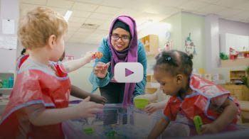 An early childhood educator is smiling as they hold a toy bunny up to a child at a watering table. The child is holding up a toy fish as they look at the early childhood educator. 