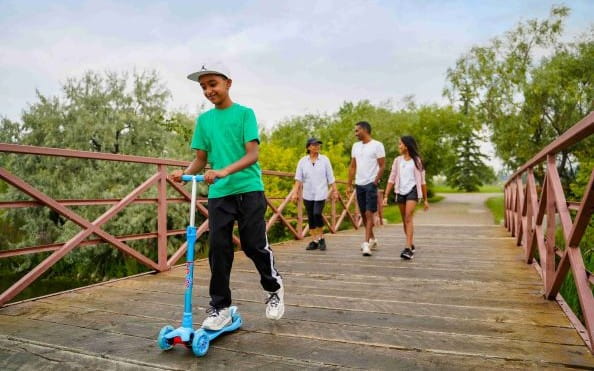 A child in a green shirt rides a blue scooter across a wooden bridge while his parents and sibling walk behind