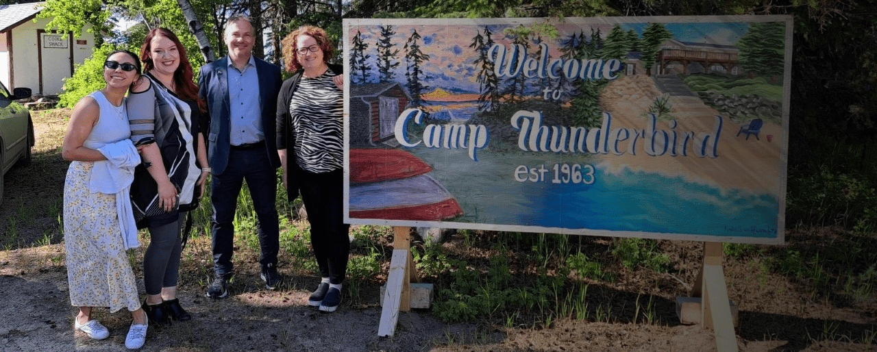 Group of people standing outdoors beside a large painted sign that reads ‘Welcome to Camp Thunderbird est. 1963,’ featuring a scenic lakeside illustration with trees, boats, and cabins.
