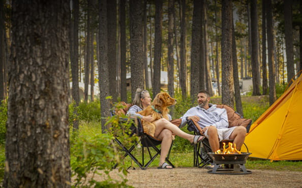 A person sitting on a chair with a dog on her lap is talking to another person sitting in a chair in front of a campfire and beside a tent at a campsite the is surrounded by trees.