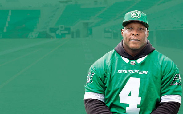 Micah Johnson, a defensive lineman with the Saskatchewan Roughriders, wears a retro Riders logo hat and green jersey with the inside of Mosaic Stadium in the background.