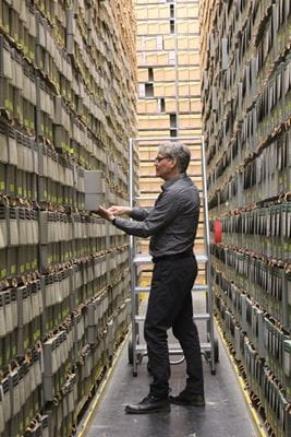 An older man wearing black pants and grey shirt stands in narrow corridor between 2 two-metre high shelving units filled with files, books and papers at the Provincial Archives of Saskatchewan.