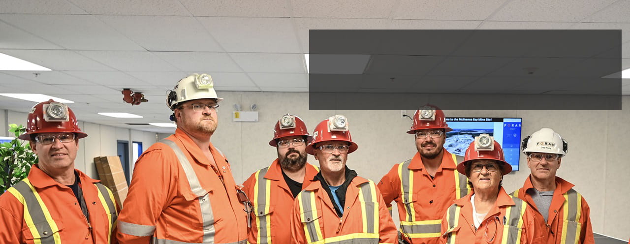 A group of 7 members from the Northern Saskatchewan Environmental Quality Committee, wearing orange coveralls with reflective stripes and hard hats fitted with lights, stand in a row in a conference room in northern Saskatchewan