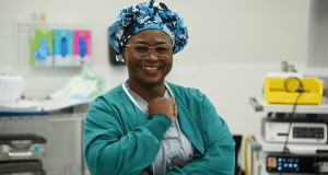 Dr. Mofolashadé Onaolapo, Anesthesiologist with the Saskatchewan Health Authority, in hospital a setting, wearing medical scrubs and cap, standing and smiling. 