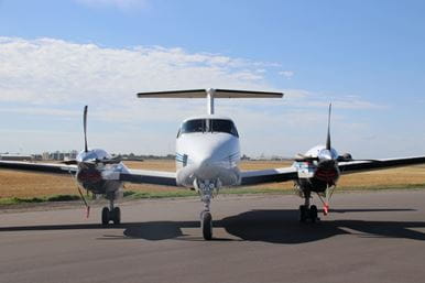 Front view of a twin-engine aircraft parked on a runway under a clear blue sky.
