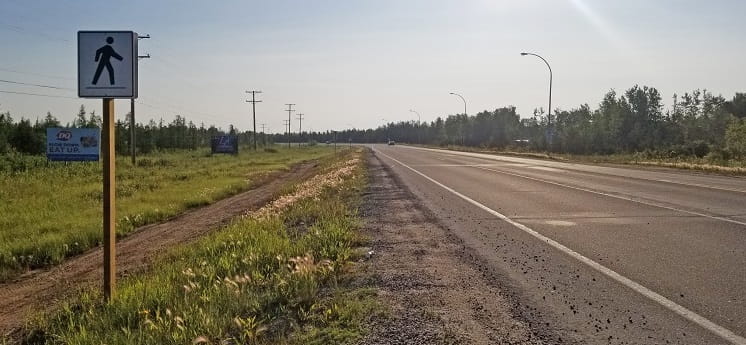 Paved highway with grassy ditch and a white and black pedestrian sign on the left side