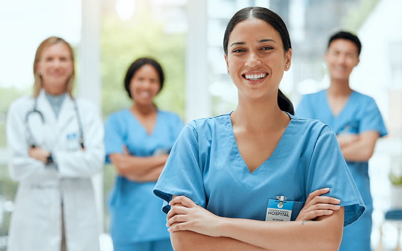 Female nurse wearing scrubs while standing with her arms crossed infront of three colleagues behind her. 