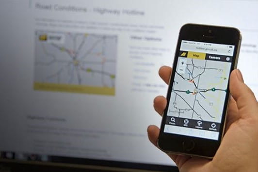Hand holding mobile device with computer desktop in background. Both devices show the Highway Hotline map.