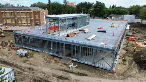 A steel-framed building under construction with an open upper floor, surrounded by soil, equipment and cones.