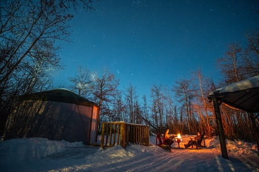 A round yurt-style tent sits on the left, partially surrounded by snow, with a small wooden deck in front and a campfire to the right.