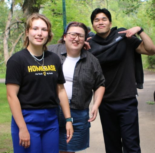 Two young women and one young man show their HOMEBASE t-shirts to the viewer. They are smiling and standing in a park during the summer.