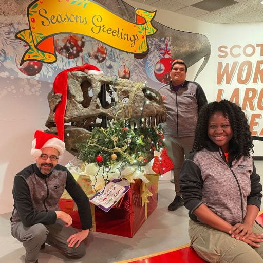 Staff of Royal Saskatchewan Museum pose with a T.rex skull with a decorated Christmas tree in his mouth being used as a holiday themed display. A yellow sign behind reads Seasons Greetings. The display is part of the holiday season programing at the museum.