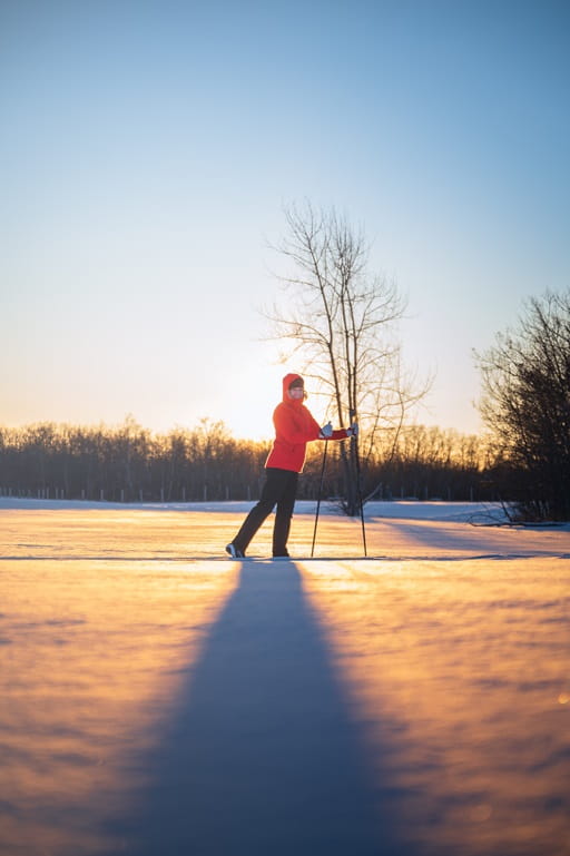 A person in a red jacket cross-country skiing on a snow-covered field during sunset.