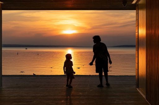 An adult and a child stand in shadow, facing a sunset on Waskesiu Lake in Saskatchewan. Birds can be seen flying over, and resting on the surface of the water. This image, taken by Brenda Makin, is one of the winners of the 2024 ExploreSask Photo Contest. 