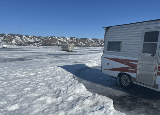A camper trailer resting on ice during winter, set against a backdrop of blue skies and snow, with another trailer afar. 