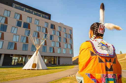 Dakota Dunes Resort - A woman, wearing traditional Indigenous regalia, stands in the foreground with her back to the camera. The Dakota Dunes Resort is pictured in the background of the image, with a tipi set up on the grass in front of the resort. Dakota Dunes Resort won the Tourism Employer of the Year award at the Canadian Tourism Awards on Thursday, Nov. 20, 2025.