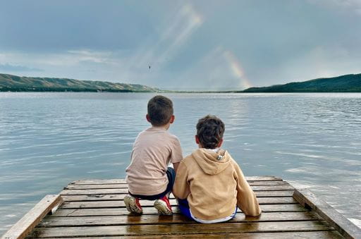 Twin Shores Lodging, Bird's Point, SK - Lindsey MacLeod - 2025 Grand Prize Winner Two kids sit on a dock looking out towards a lake at Twin Shores Lodging in Bird's Point, Saskatchewan. This image, taken by Lindsey MacLeod, won the grand prize of the 2025 ExploreSask Photo Contest.