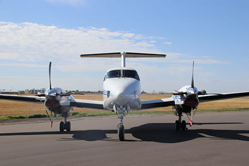 A white airplane facing forward with two propellers on a runway with new black pavement. The background is a blue sky over a farmer's field.