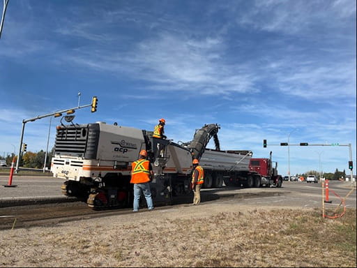 Heavy equipment with tracks dumps road material into a nearby semi truck to help build a turning lane in the Battlefords near an intersection with street lights. Construction workers are nearby in orange hats and safety vests. The sky is a bright blue with some clouds.