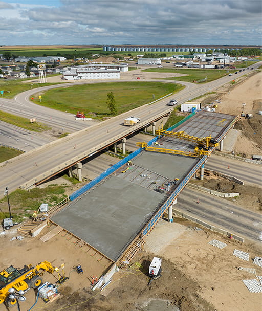A concrete and steel highway overpass under construction with various equipment nearby facing the edge of Moose Jaw. A blue prairie sky is in the background on a clear day.