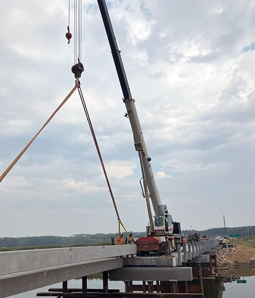 A crane lifts concrete over a bridge in mid construction over a river with clear and still water with trucks and construction workers dressed in orange safety gear nearby. The scene is in northern Saskatchewan with a forest surrounding the area. The sky is overcast.
