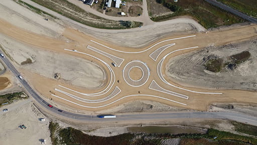 An aerial view of the Highway 39 roundabout under construction at Weyburn. Concrete circles have roads approaching the shape. Most of the surfaces are covered with tan-coloured sand and gravel.
