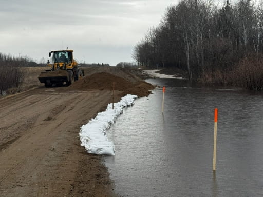 A rural road beside a swollen river is partially flooded. Sandbags line the road’s edge to hold back water, while a front-end loader drives along the muddy roadway. Leafless trees stand on the right, and water covers the adjacent ditch and low-lying land.