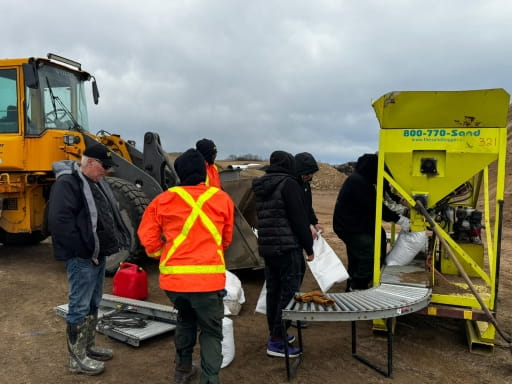 A group of workers in jackets and safety gear operate a sandbagging machine next to a large front-end loader. They fill and handle white sandbags on a conveyor system, preparing flood protection materials on a muddy roadside.