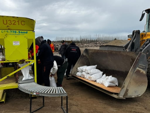 Workers continue filling sandbags using a machine and conveyor, stacking completed bags inside the bucket of a front-end loader. The surrounding area appears wet and muddy, with more equipment and sand piles in the background.