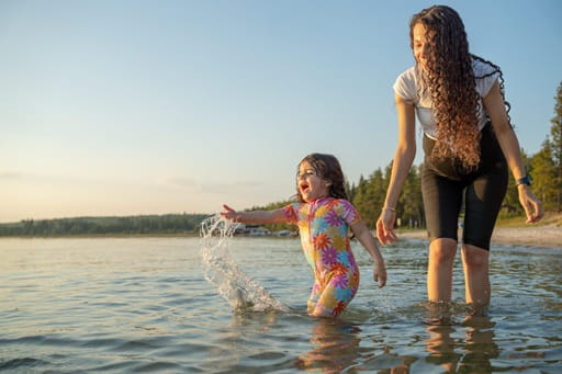 An adult and a young child stand in shallow lake water at sunset, with the child splashing water while the adult looks on near the shoreline, trees visible in the background.