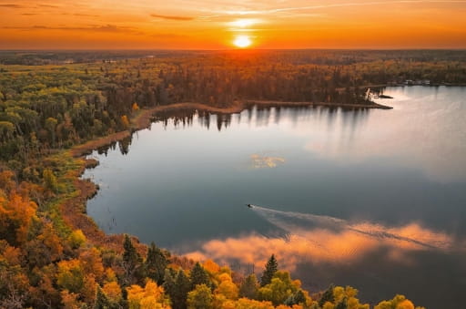 An aerial view of Christopher Lake. The sun sits on the horizon in the background, casting an orange glow in the sky. Trees surround the lake in various shades of green, yellow, and orange.