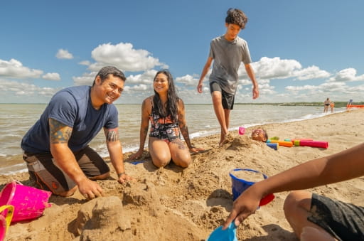A family plays in the sand at Good Spirit Lake Provincial Park. Pictured behind the family is the lake, and a blue sky with scattered clouds. A group of people are seen playing in the water in the background.
