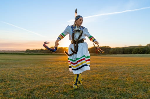 A woman, wearing traditional Indigenous regalia, dances in the grass at Fort Carlton Provincial Historic Park. The sun shines a golden glow on the horizon from the background.