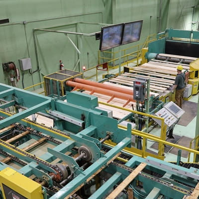 Industrial lumber processing equipment operates inside a sawmill facility. Conveyor systems, rollers, control panels, overhead monitors, and large machinery are visible, while a worker wearing a hard hat stands near the equipment monitoring operations.