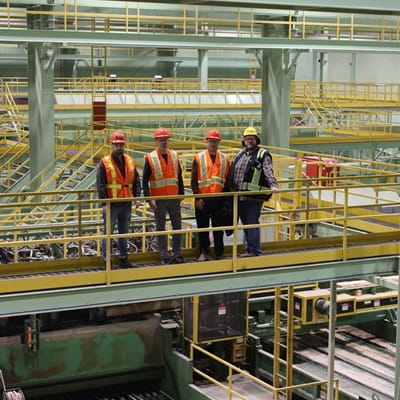 Four workers wearing orange reflective vests and hard hats stand together on a catwalk inside a sawmill or industrial processing plant. The large facility contains interconnected walkways, railings, machinery, and conveyor systems extending throughout the background.