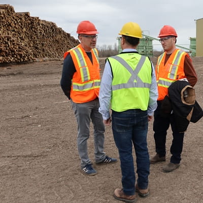 Four workers in reflective safety vests and hard hats stand on an elevated walkway inside a large industrial lumber processing facility. Yellow railings, metal platforms, and heavy machinery surround them across multiple levels of the plant.
