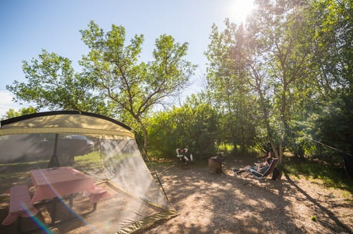 Three people sit around a campfire pit in Danielson Provincial Park on a sunny day. There is a screened tent around a picnic table.