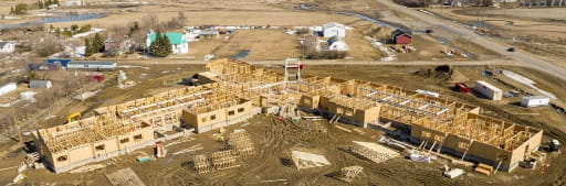 Aerial view of the Grenfell long-term care home under construction, showing the wooden framing of the building across a large rural construction site with surrounding fields, roads, and nearby homes in early spring conditions