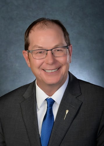 Picture of MLA Keisig in a dark blue suit with a MLA pin on the lapel, a white shirt with royal blue tie. He has dark brown hair and wears wire-rimmed glasses. 
